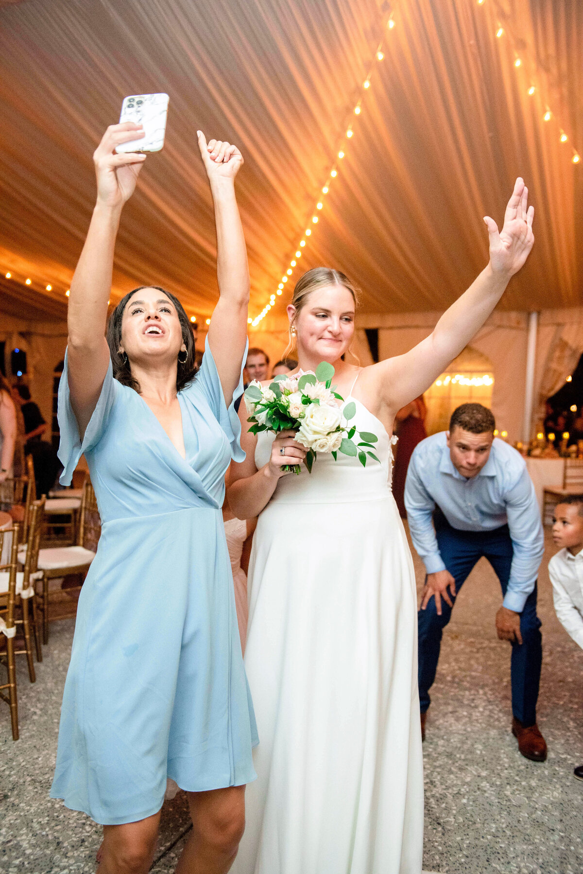 Two women celebrate at a wedding. One in light blue raises a phone and her arm, while the other in a white dress holds flowers and raises her arm. String lights decorate the tent. People are visible enjoying the event in the background.