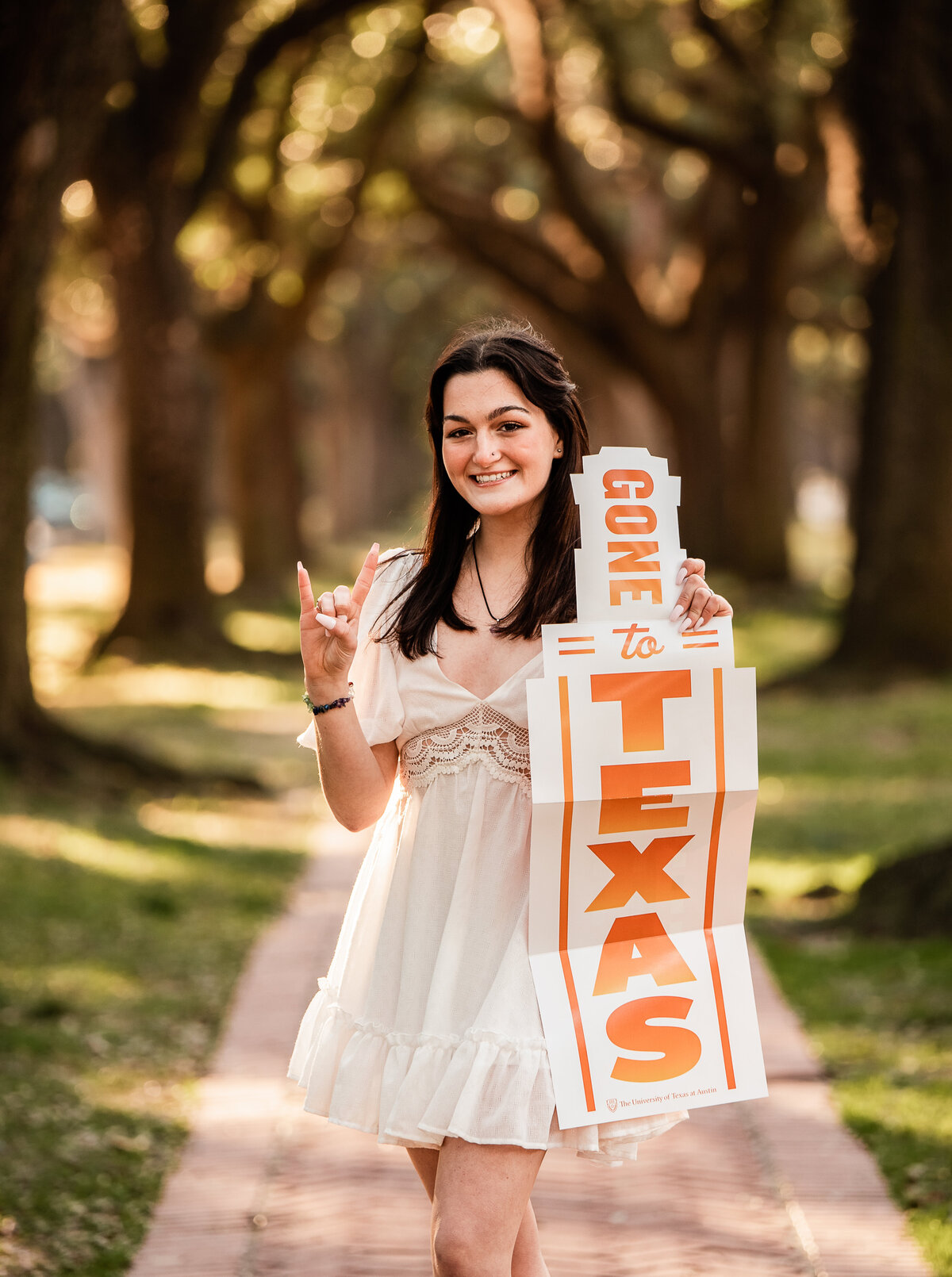 A graduate heading to University of Texas in the fall stands on a tree lined path holding a UT banner.
