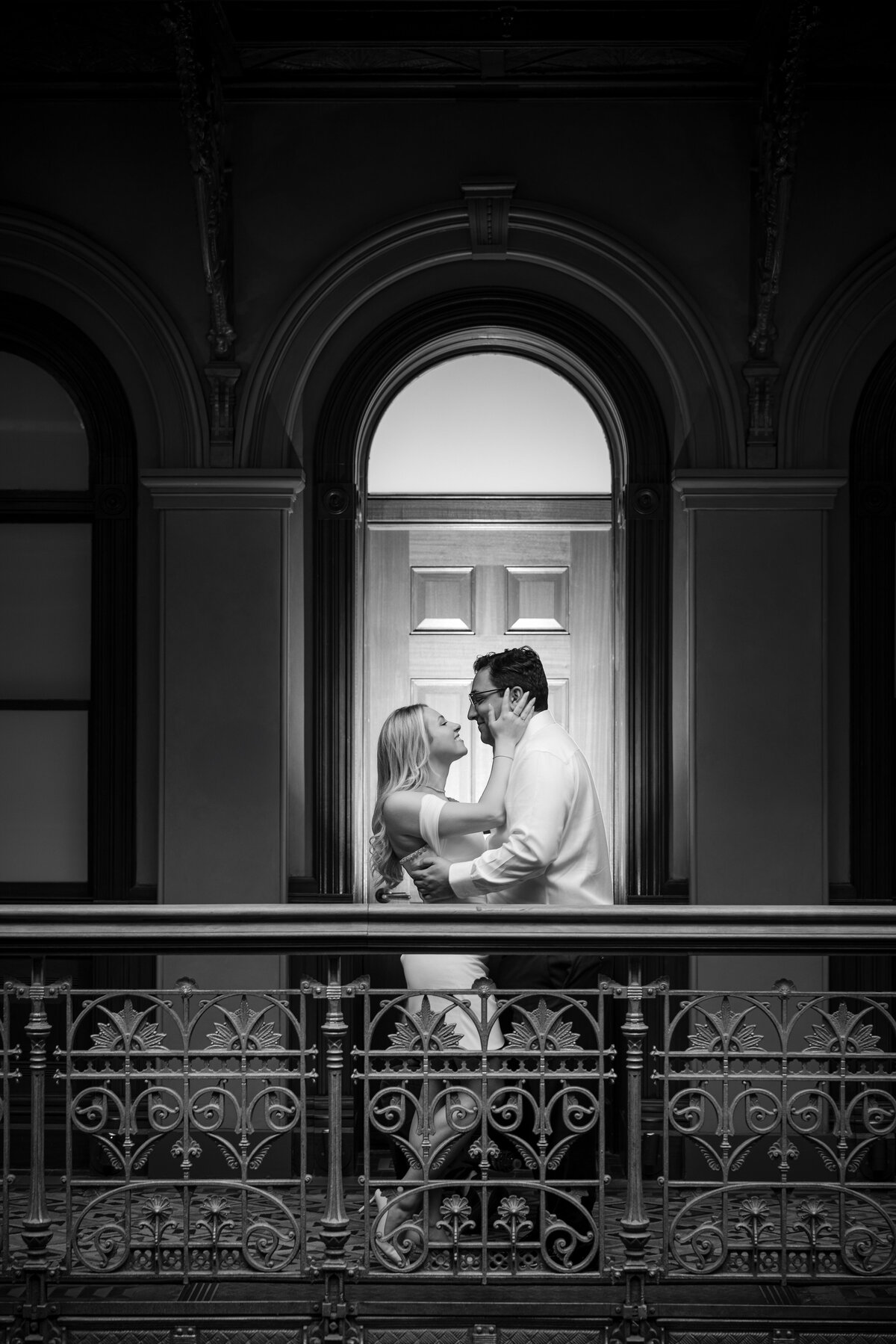A black and white image of a couple framed symmetrically in front of an arched window and paneled door, sharing a quiet moment on the ironwork balcony of the Beekman Hotel—an example of editorial Beekman Hotel engagement photos that highlights both architectural elegance and intimate connection.
