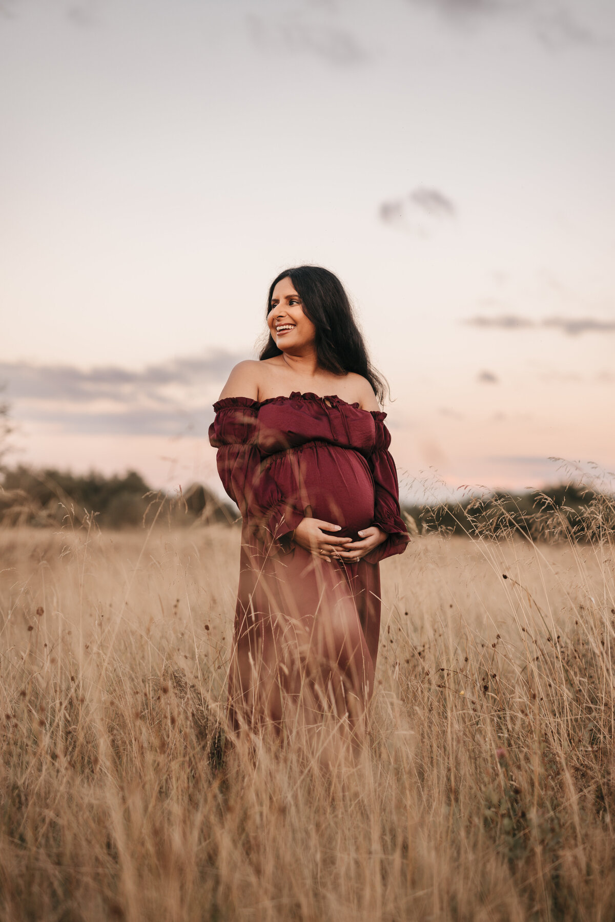 Photo of a pregnant woman standing in a red dress amongst long grass at sunset
