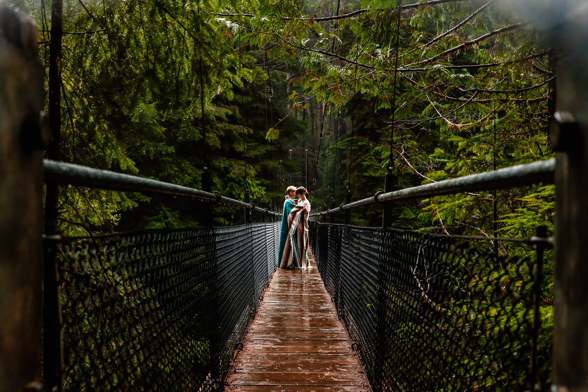 wrapped in a blanket, two brides snuggle on a bridge in the middle of a moody forest duiring their washington elopement