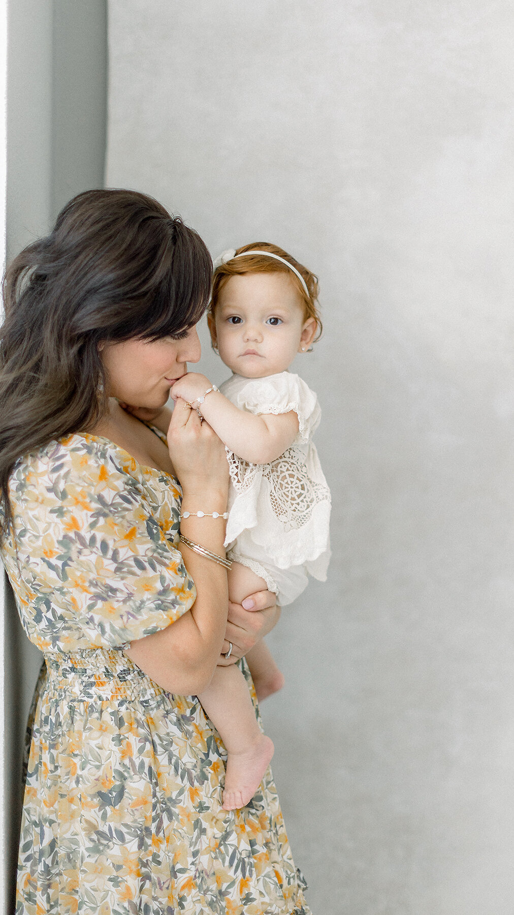 A mother standing in a DFW Photography studio while she is holding her baby and kissing her hand.