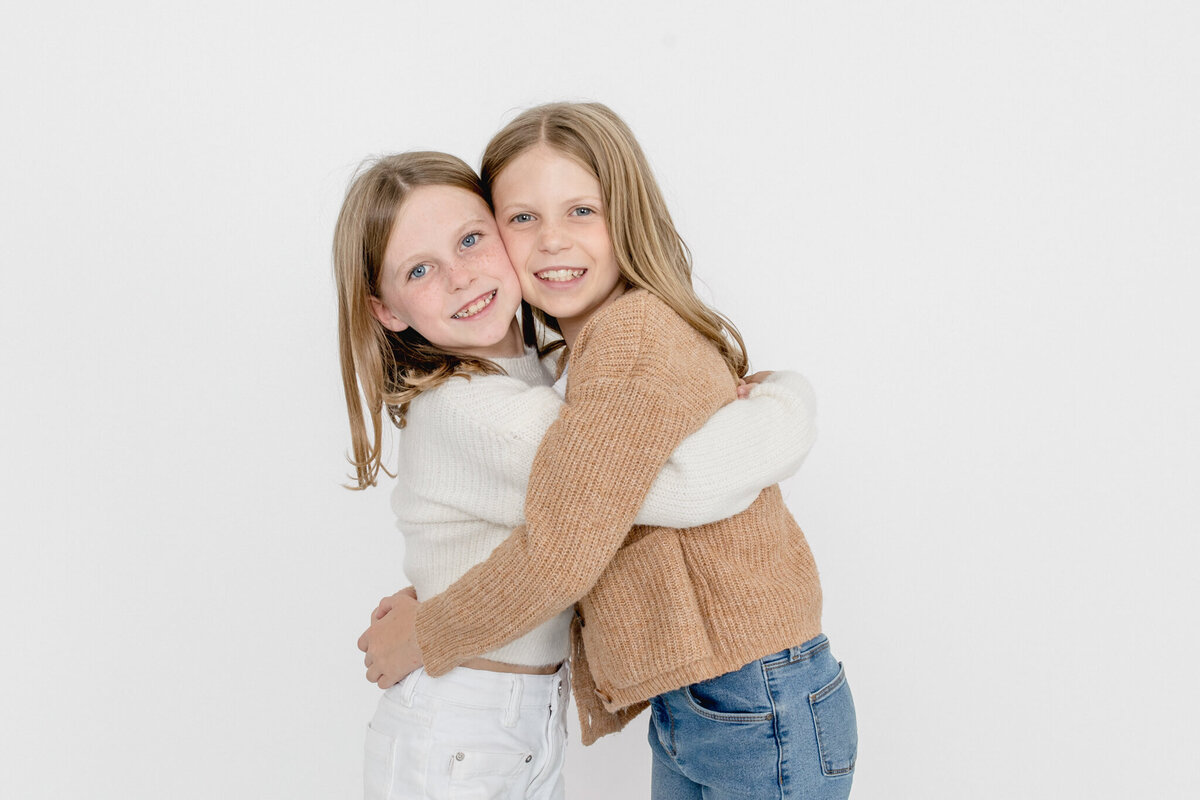 Two young sisters with blonde hair, hugging each other with their heads facing the camera and their cheeks pressed together, smiling against a white background.