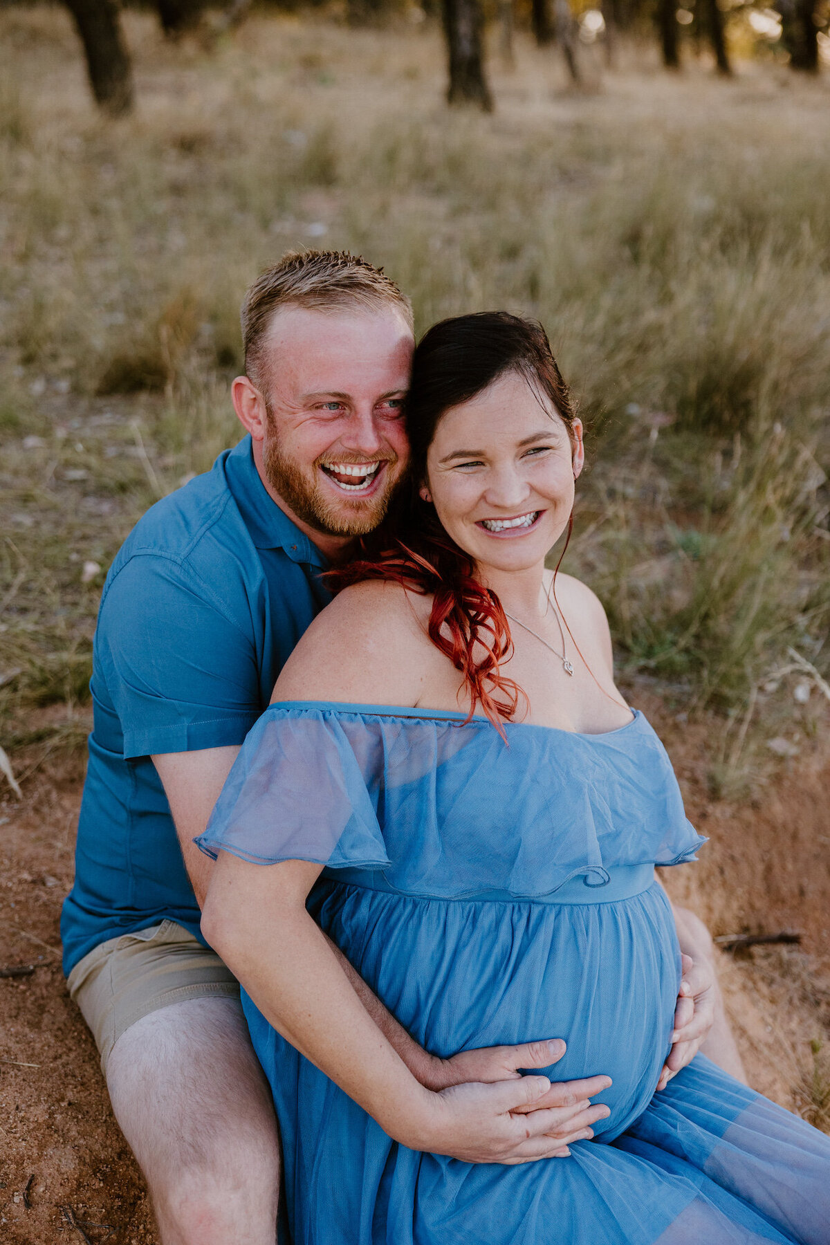 Loved-up couple embracing, with her partner snuggling into her neck from behind and laughing while holding her belly during a relaxed maternity session.