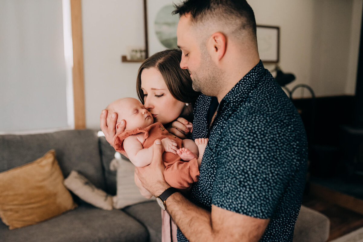 A young couple hold their newborn daughter together in their living room while the mother gives her a sweet kiss on the cheek