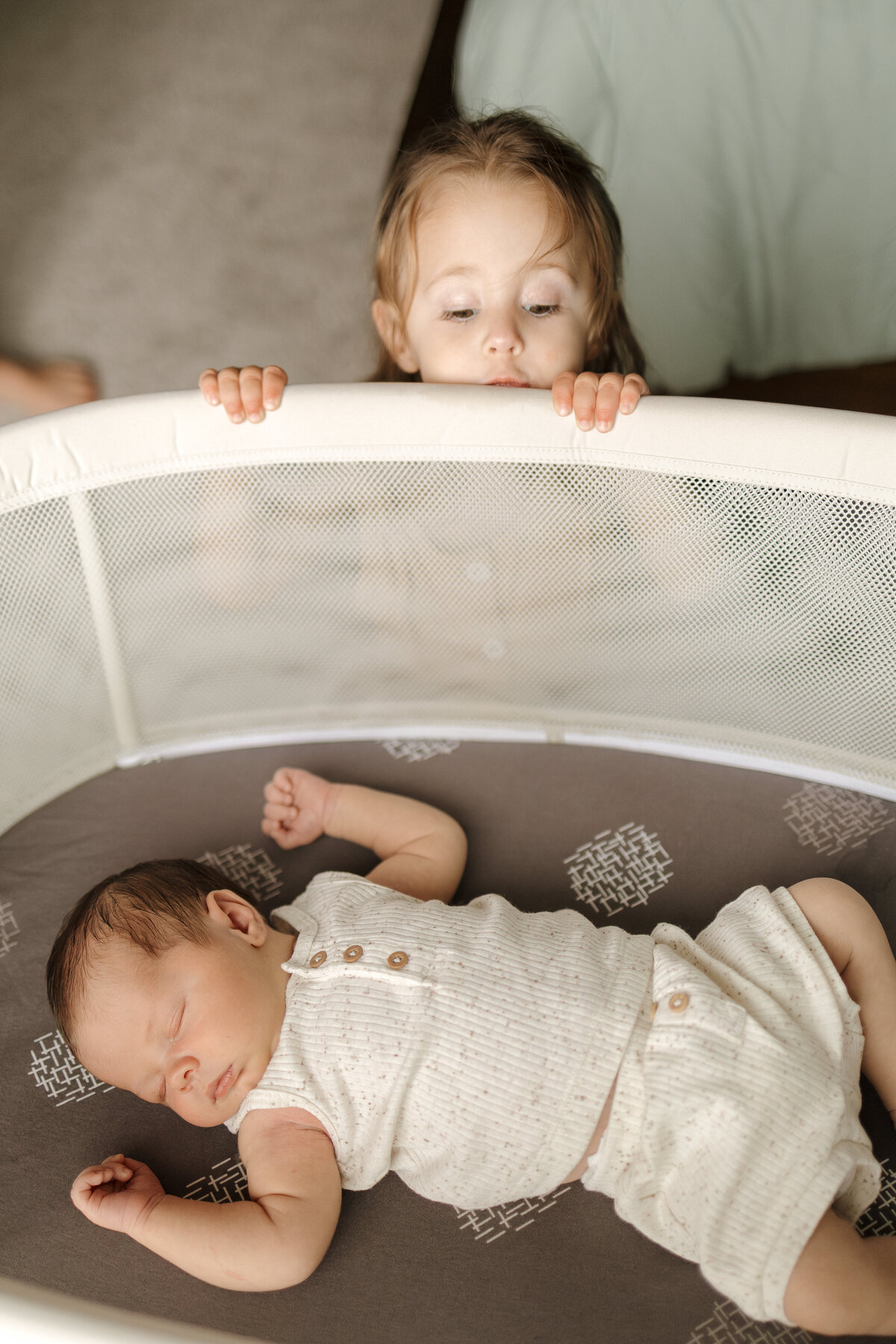 big sister peeping over bassinet as newborn baby brother is sleeping during lifestyle in home newborn session in longmont colorado with alexis adkins photography