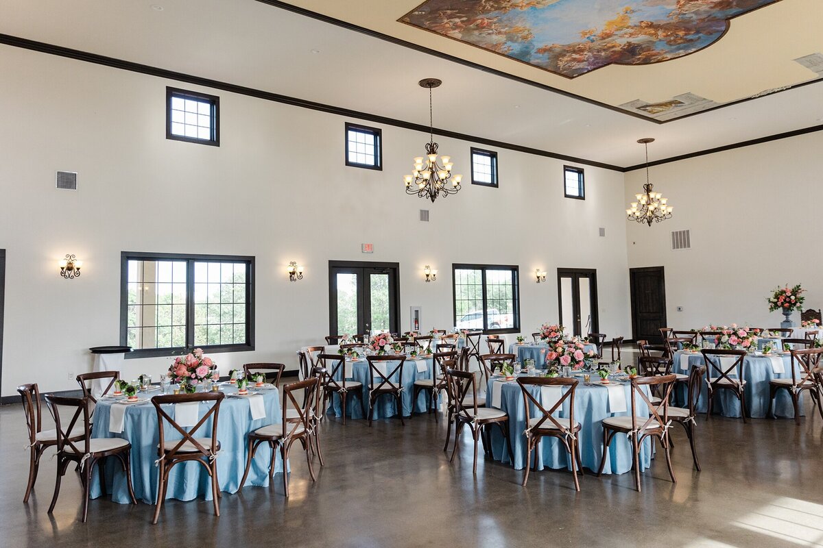 A room shot of a wedding reception at Stoney Ridge Villa in Azle, Texas. The large white room has multiple chandeliers hanging from the ceiling, a large intricate mural painted on the ceiling, and many receptions tables around the room. each reception table is draped with a long blue tablecloth and has multiple place settings and floral centerpieces on it. Each table is surrounded by cushioned, wooden dining chairs.