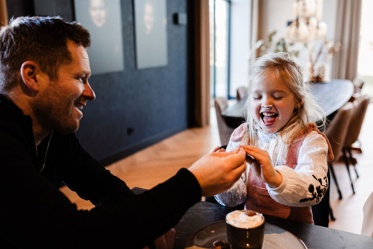 Papa en dochter zitten aan hoge tafel met warme chocomel met slagroom en papa drukt slagroom tegen neus dochter