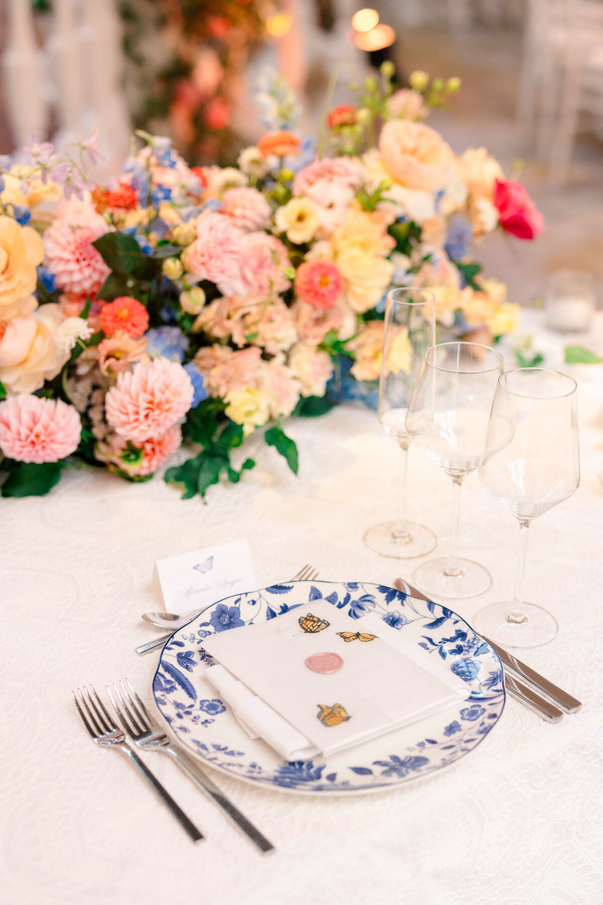A beautifully set table with a floral centerpiece of assorted colorful flowers. A blue and white patterned plate is arranged with a folded napkin displaying whimsical fruit and butterfly illustrations, accompanied by three wine glasses and silverware.
