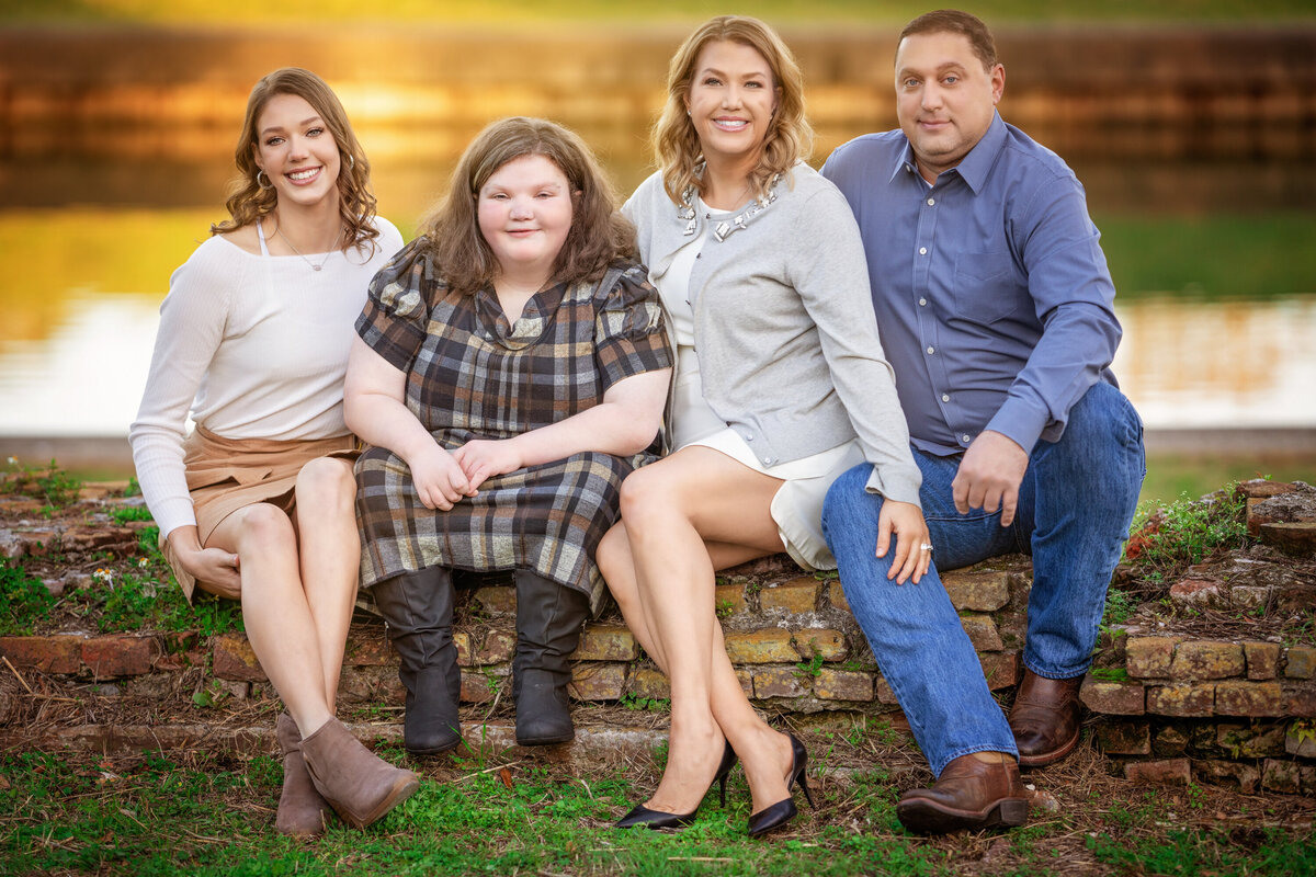 Mom, Dad, and 2 daughters are sitting in front of a canal in New Orleans.  One of the daughters has down syndrome.  They are all smiling and looking at the camera.