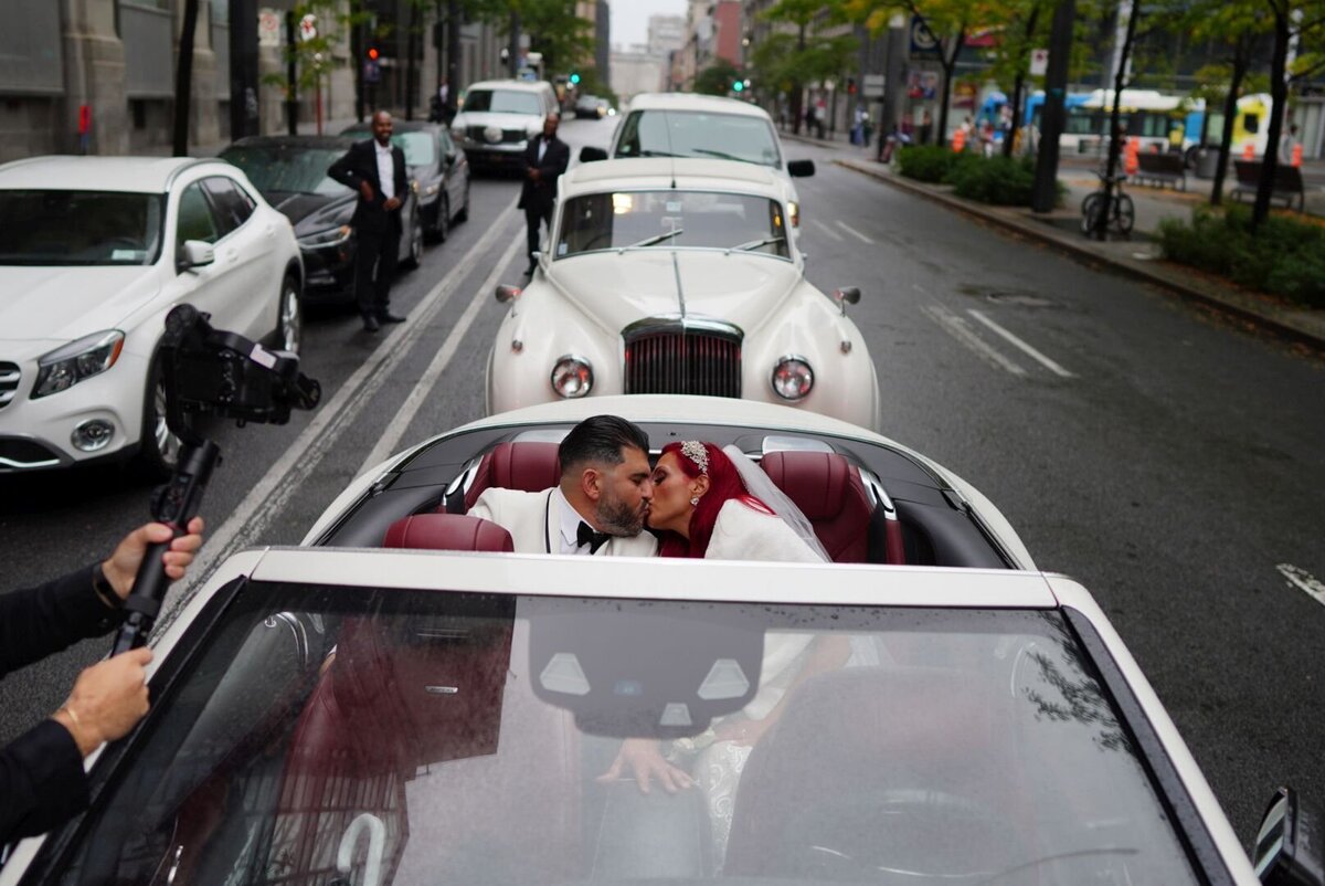 A couple shares a passionate kiss while sitting in a stylish convertible car. The image captures the joy and intimacy of their moment together, set against the backdrop of the open road and sunny skies.