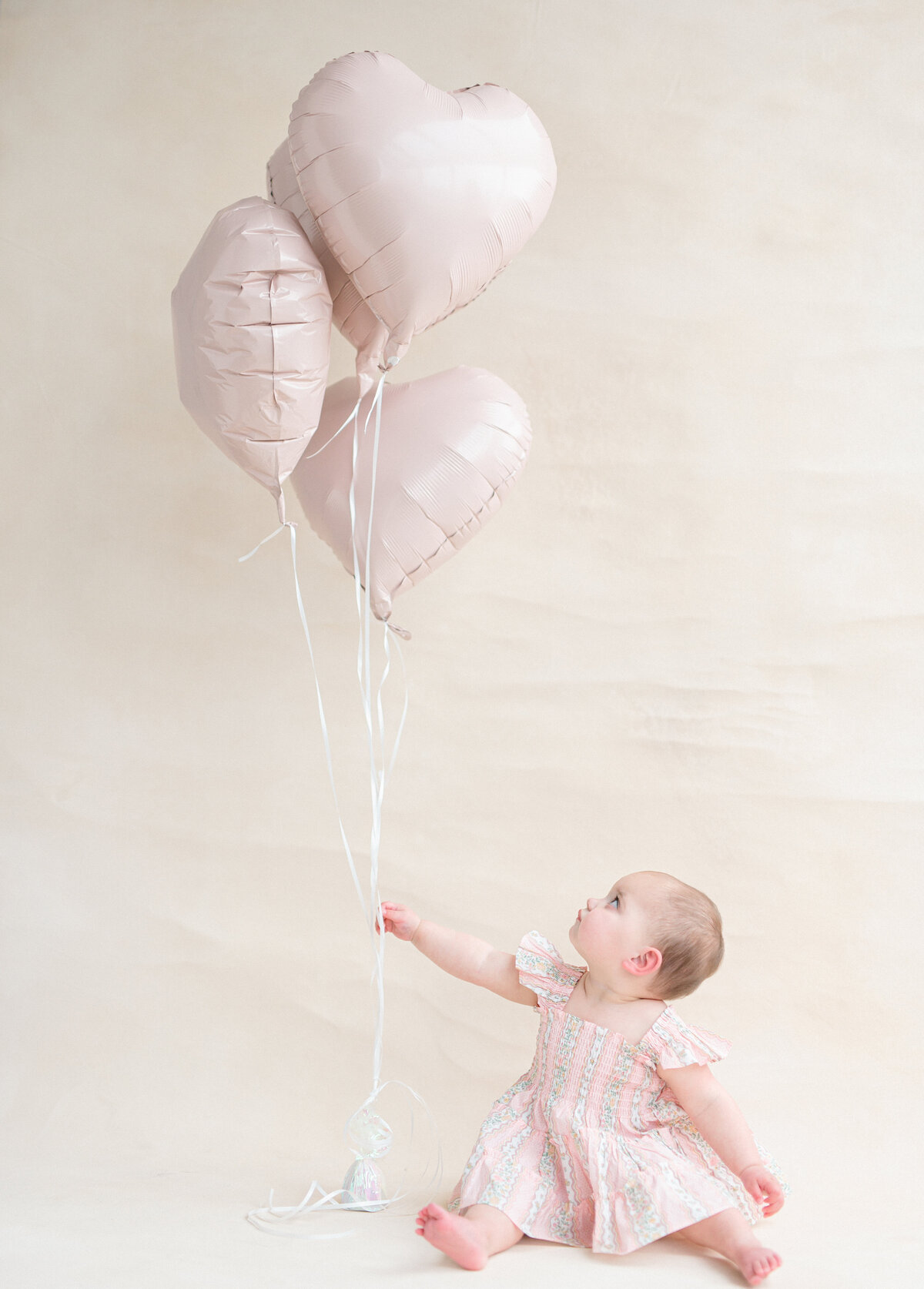 Baby girl in a pink dress looking up at heart shaped balloons 
