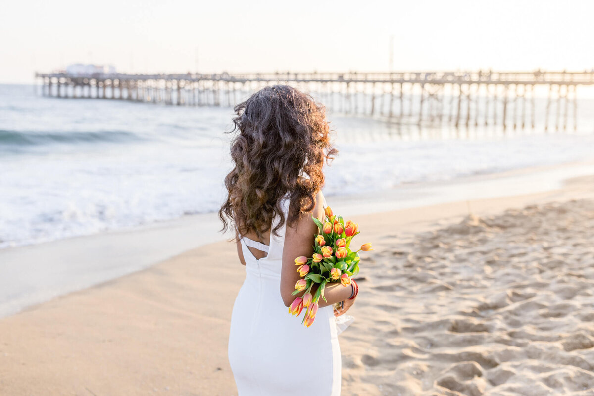 A woman in a white dress stands on a sandy beach, holding a colorful bouquet of flowers. Her back is to the camera, and she faces the ocean, with a wooden pier extending into the sea under a bright, sunny sky.