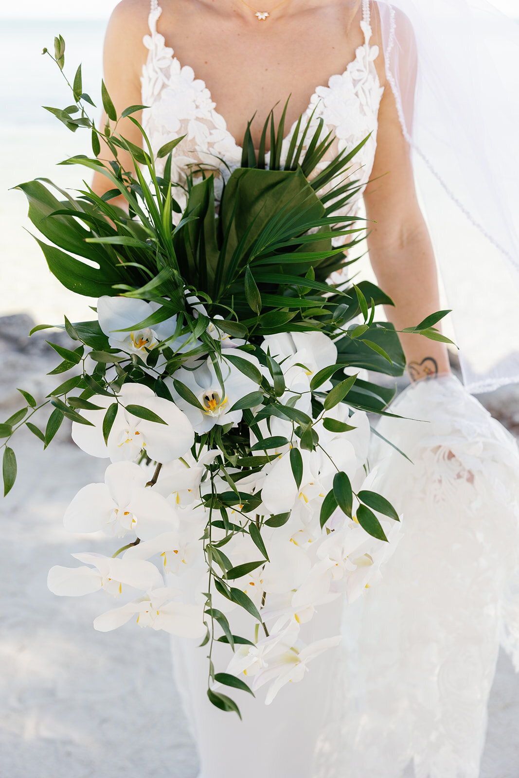A close-up of Ashley holding a cascading white orchid and greenery bouquet, with the ocean in the background.