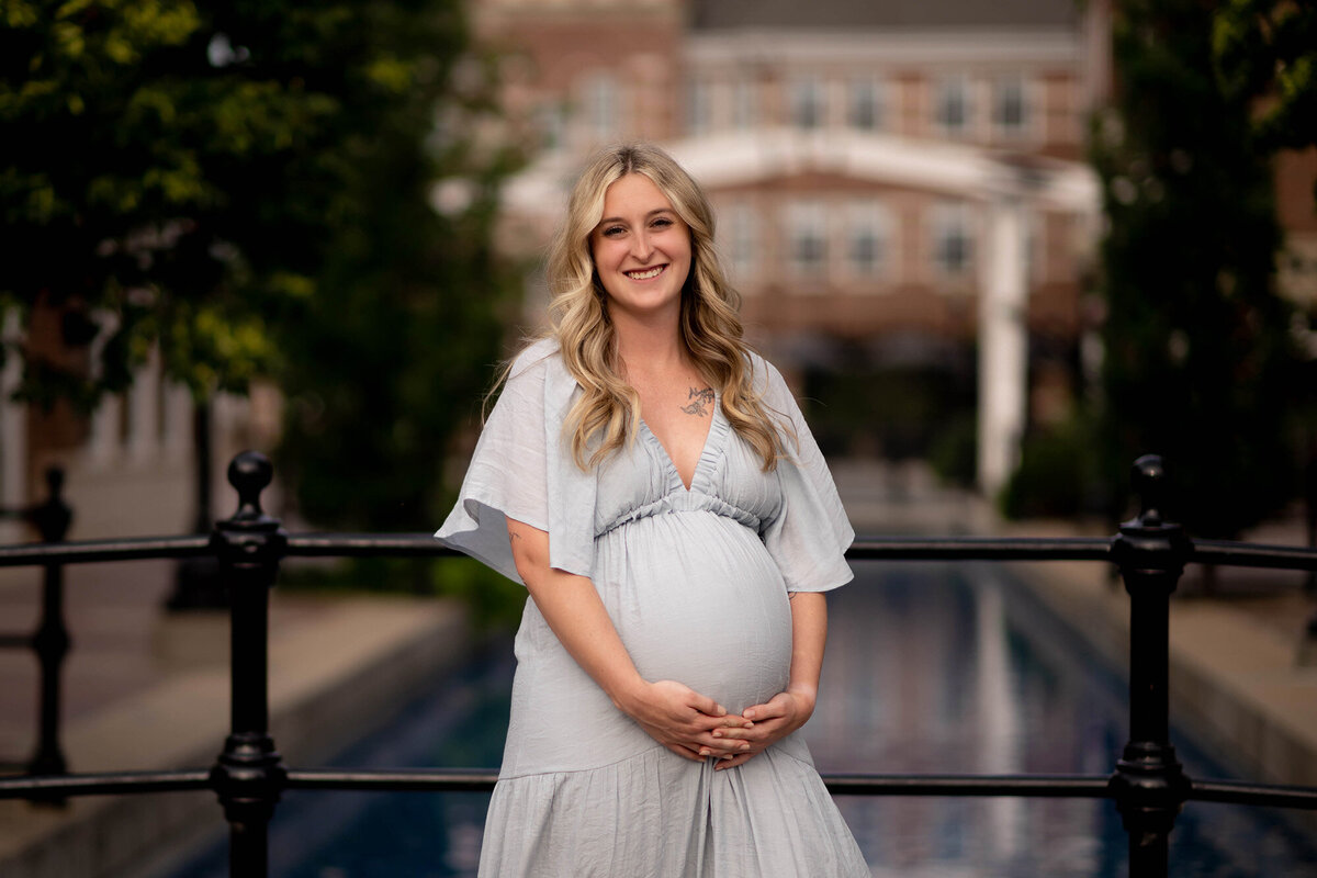 A smiling mother to be stands on a bridge holding her bump in a blue maternity gown