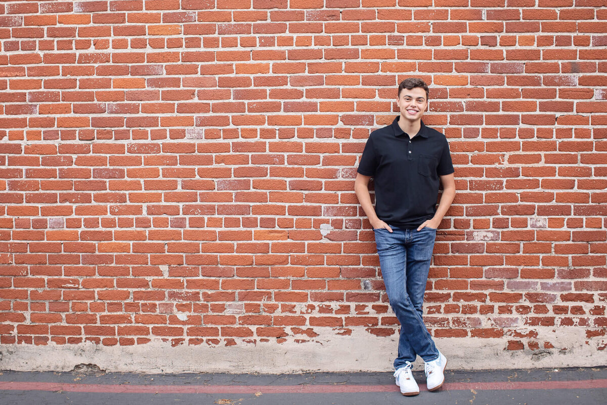 A person in a black polo shirt and jeans stands smiling against a red brick wall, with hands in pockets and wearing white sneakers.