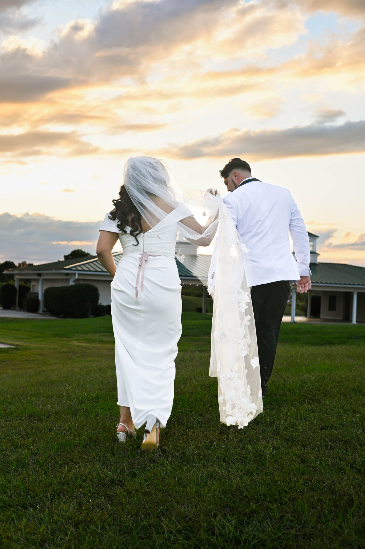 bride and groom just after wedding ceremony as they are walking up a golf course hill at their country club venue.  bride has off the should short sleeves with a mauve or rose fabric belt.  Groom in tux with white jacket and black pants.  Groom is helping bride carry her veil