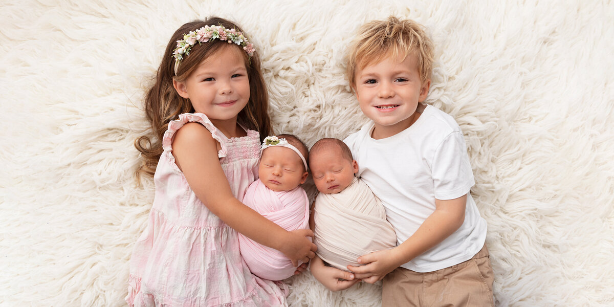 Older sister and brother sitting together holding their newborn twin siblings, smiling while the babies are wrapped and sleeping on a white fluffy blanket.
