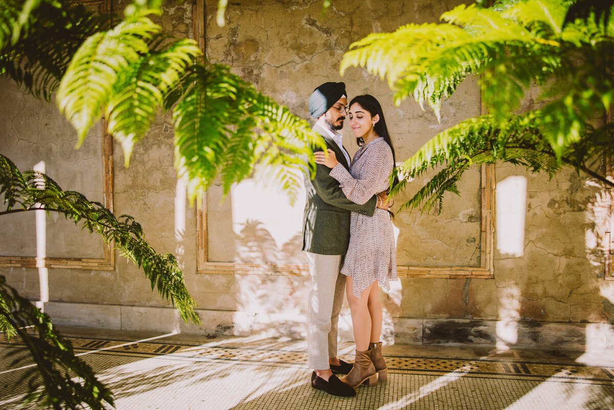 An engaged couple share a moment in front of a beige wall with plants in front at untermyer gardens in yonkers