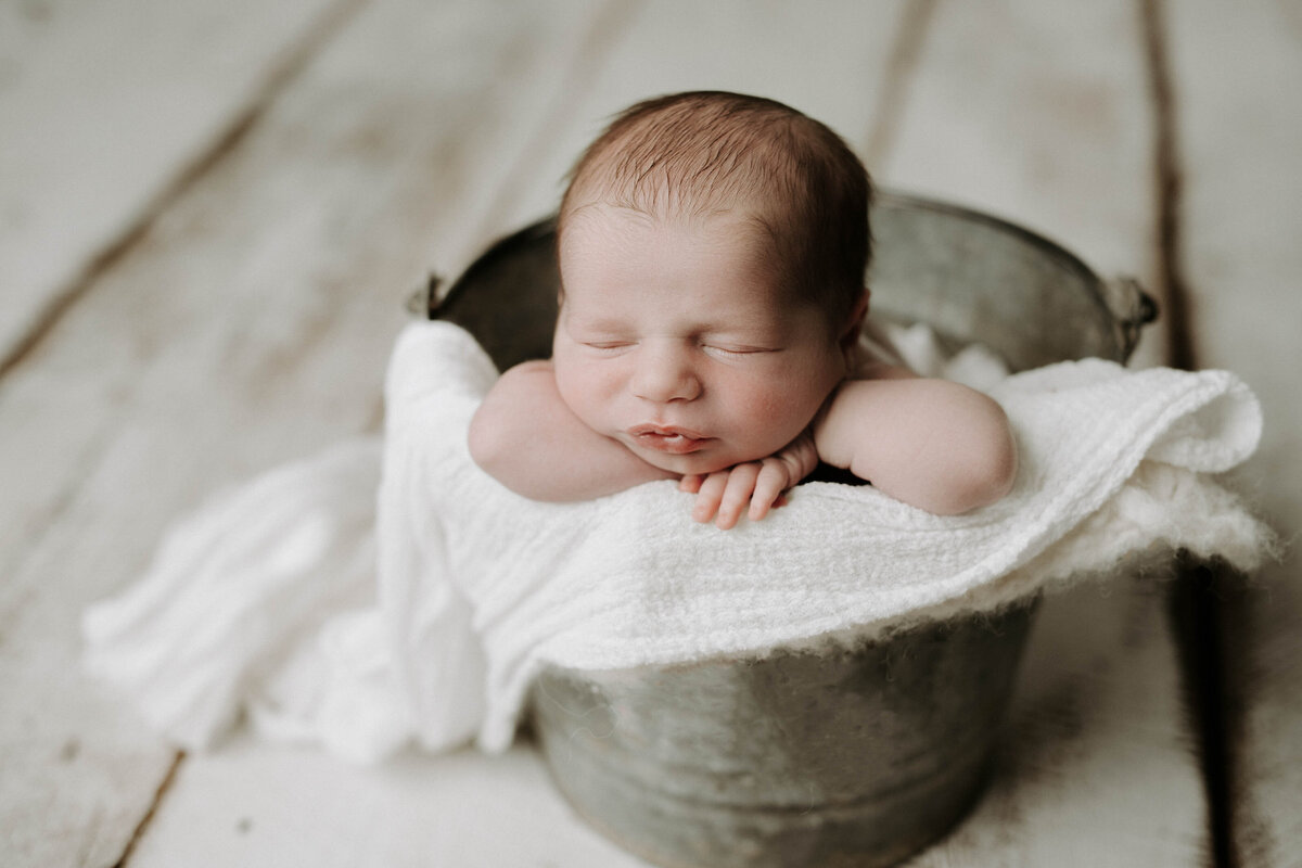 Baby in metal bucket with a white swaddle draping over the edges. Baby is resting his hands under his chin.