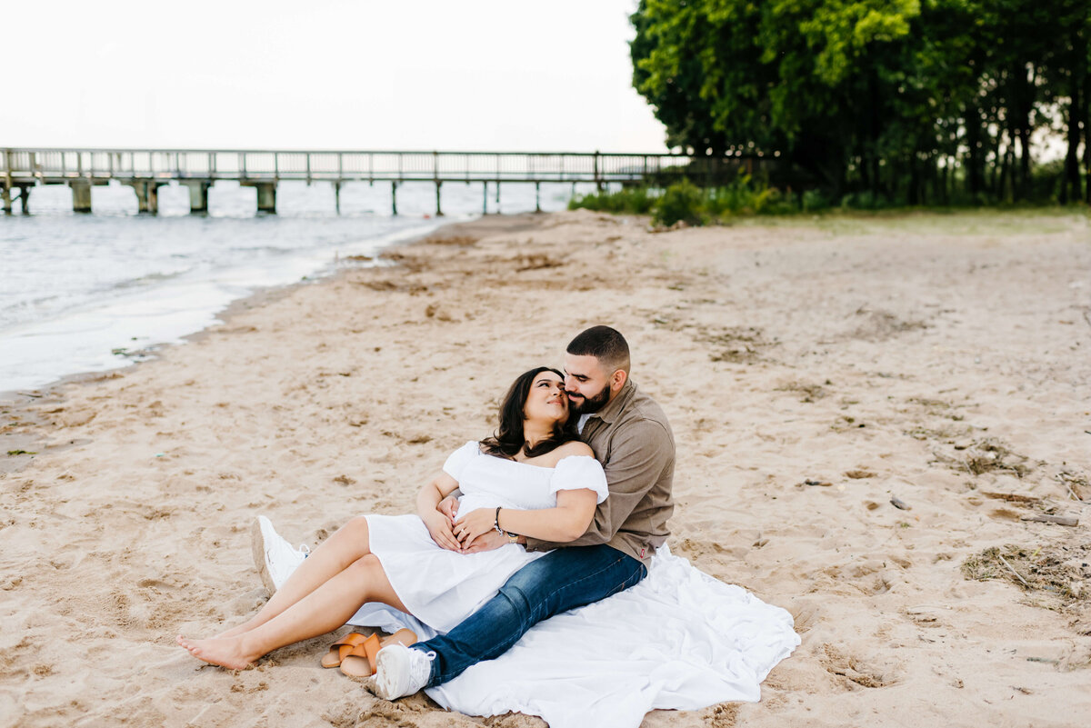 woodbridge lake couples photography with man and woman sitting together on a beach with the woman leaning into kiss the man as she smiles with a forest in the distance