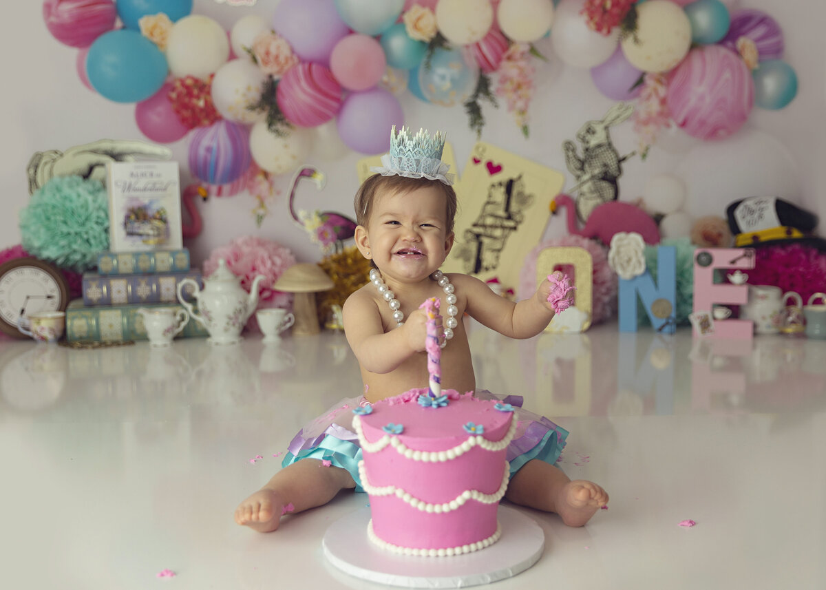 A young girl in pearls and a small crown happily sits in a New Jersey Cake Smash Photographer studio covered in cake