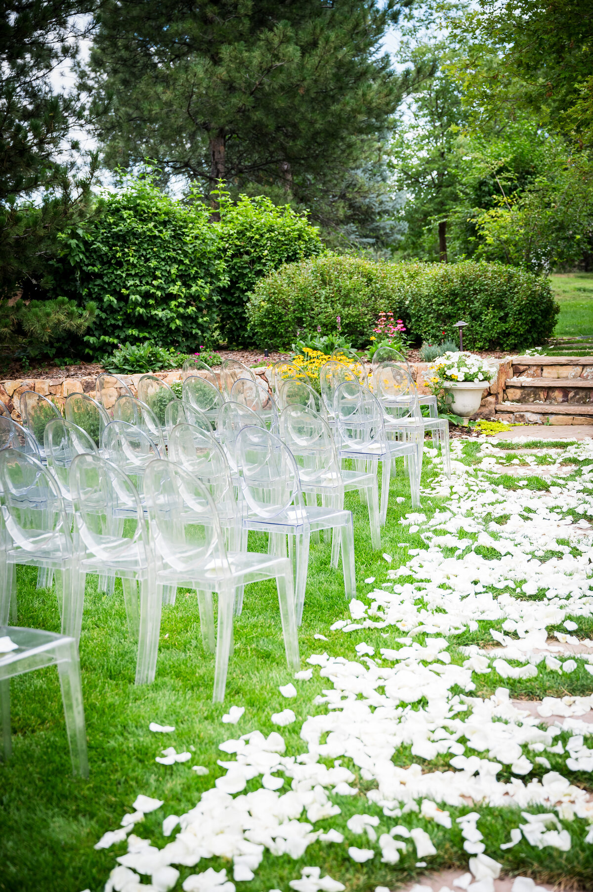 An outdoor wedding ceremony space with white flower petals all up the aisle and clear modern chairs for guests.