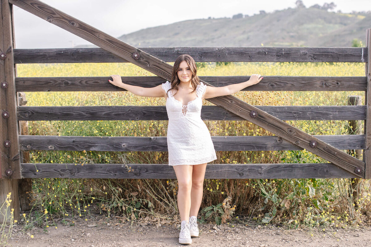 A young woman with long, brown hair wearing a short, white dress standing in front of a wooden gate with a field of grass behind her, with her arms holding onto the gate behind her on either side, looking at the camera.