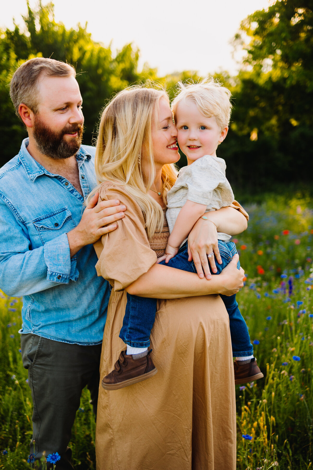 Family photograph in a garden with tall grass and small red flowers. The woman is pregnant and has a long light brown dress, she is holding her son and giving him a kiss on the cheek and he is dressed in a blue jean and a white shirt. The man has a beard and is looking at the woman in a blue outfit.