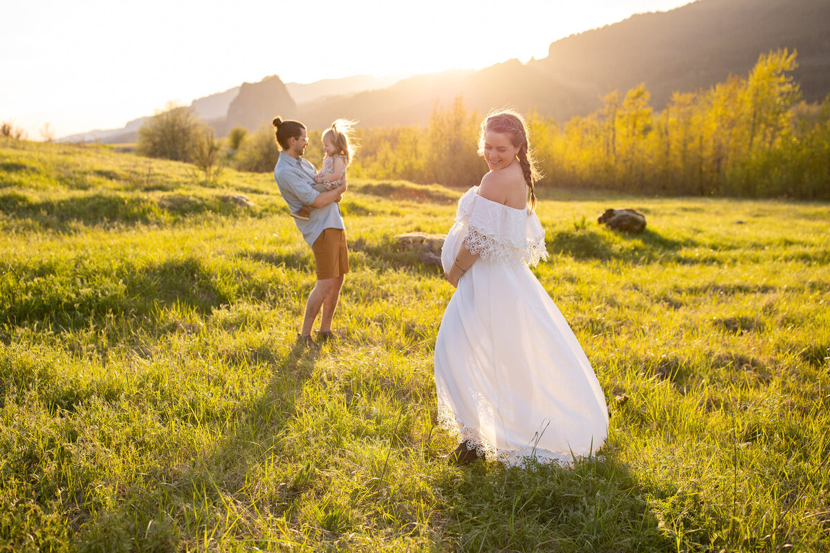 A mother standing in sunlit field holding her pregnant belly as her husband and daughter play behind her. 
