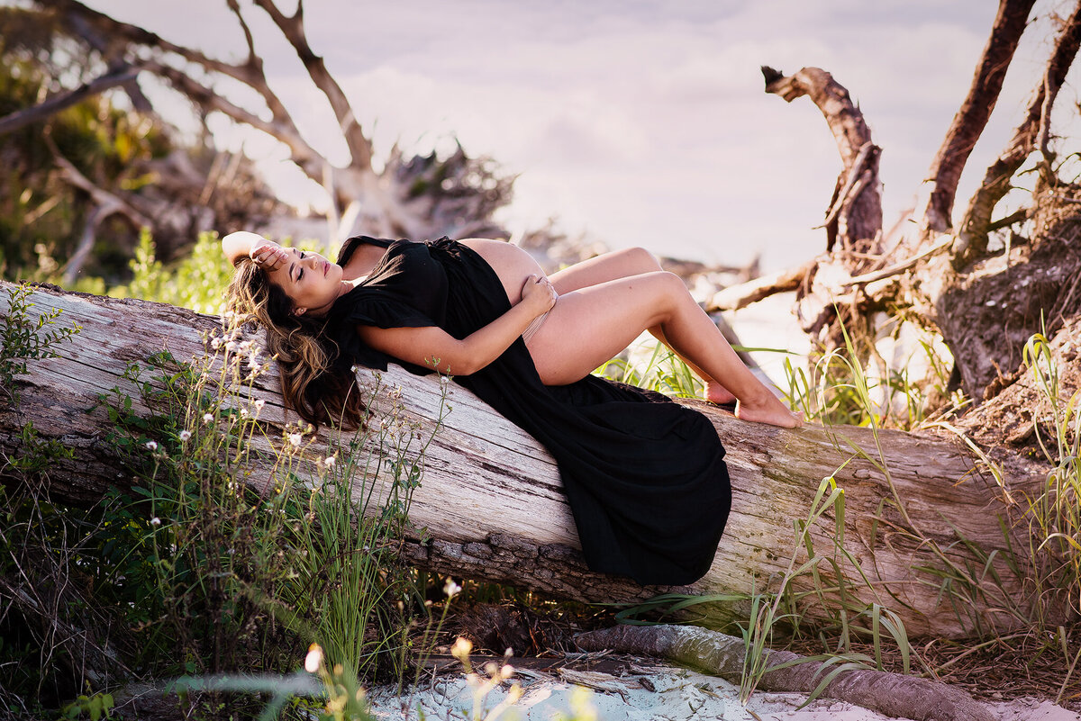 Expectant mom in black dress lying on driftwood with belly exposed at Talbot Island in Jacksonville, FL.