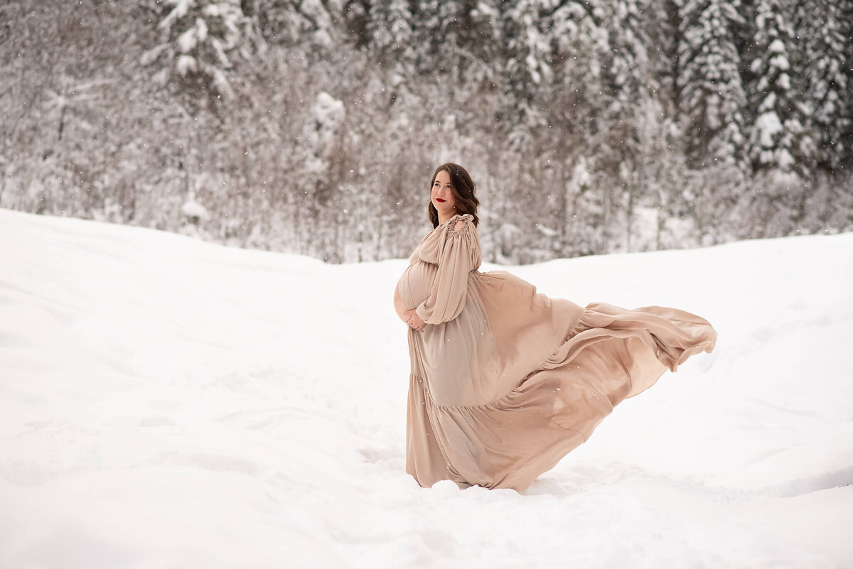 A pregnant woman wears a beige silk dress that is flowing in the wind behind her. She stands in the snow with snowy trees behind her. Captured in Sun peaks by Brittany Danielle