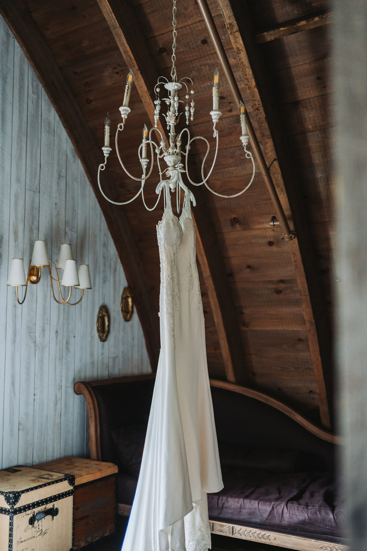 An elegant white wedding dress with intricate detailing hangs from a vintage chandelier in the rustic, high-ceilinged interior of Sweeney Barn in Manassas, Virginia, captured by Quinn Photography LLC.