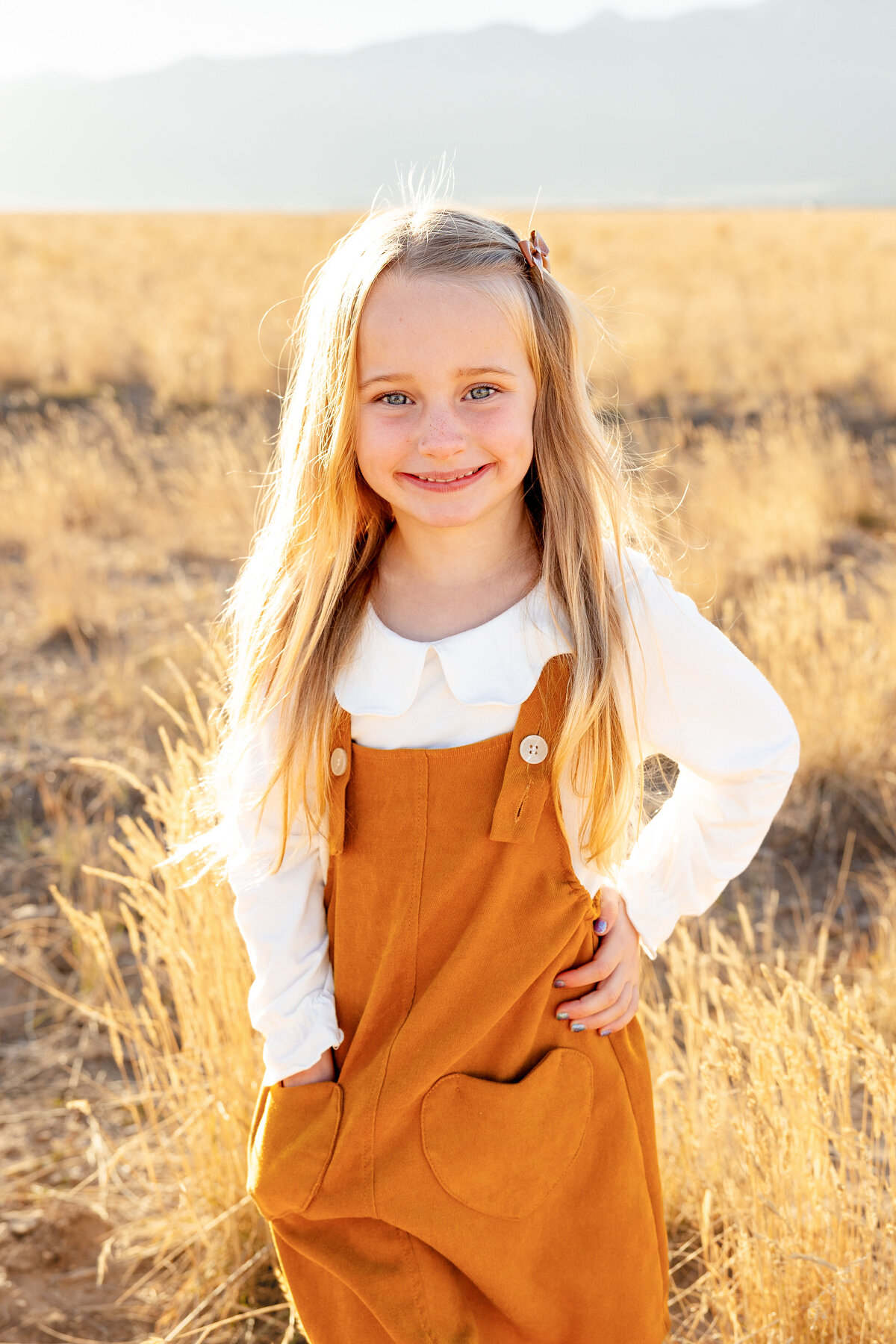 A close-up portrait of a young girl with bright blue eyes and blonde hair, wearing a mustard-colored dress in a sunlit field.