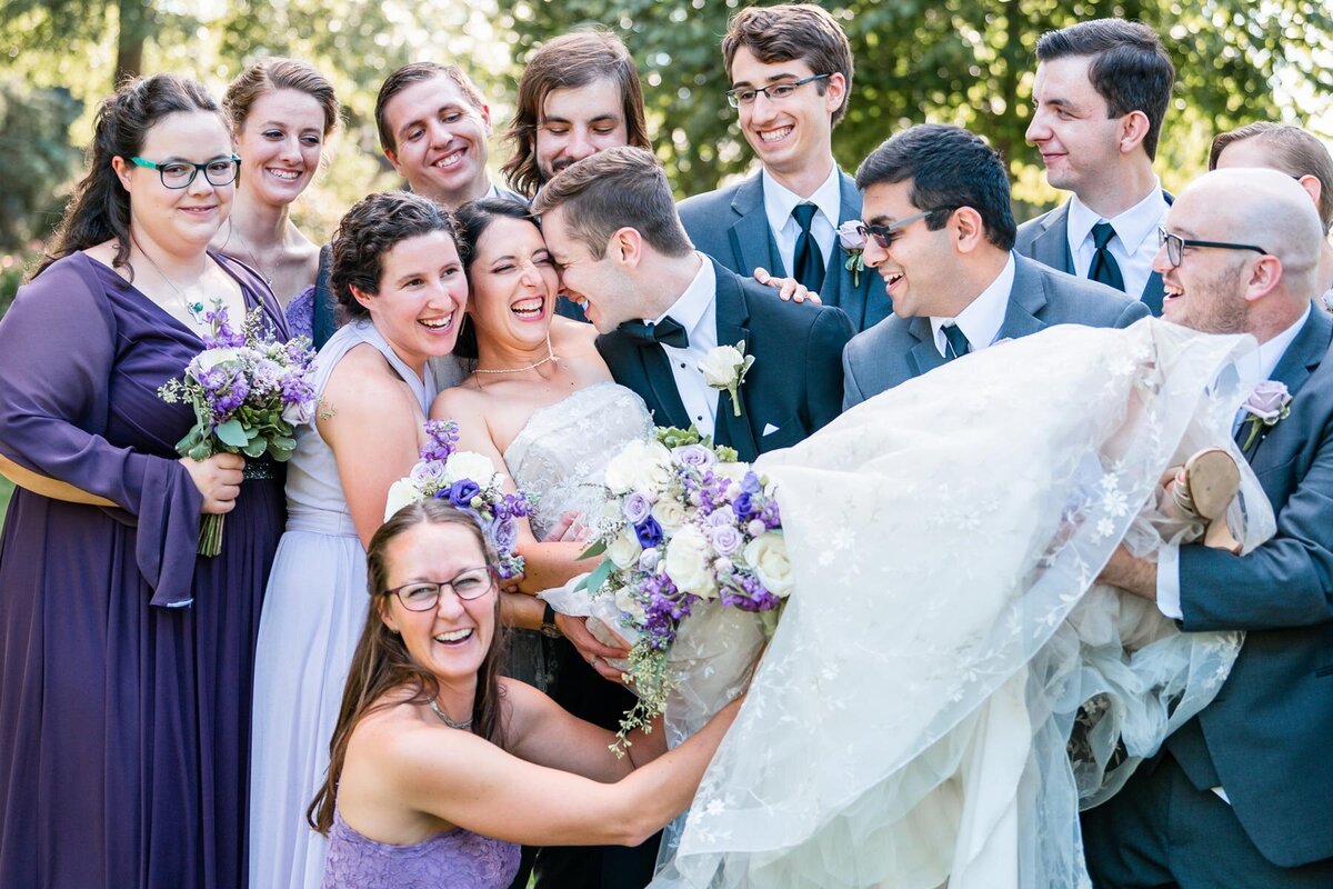 Bridal party holds bride up and laughs while groom kisses bride on cheek