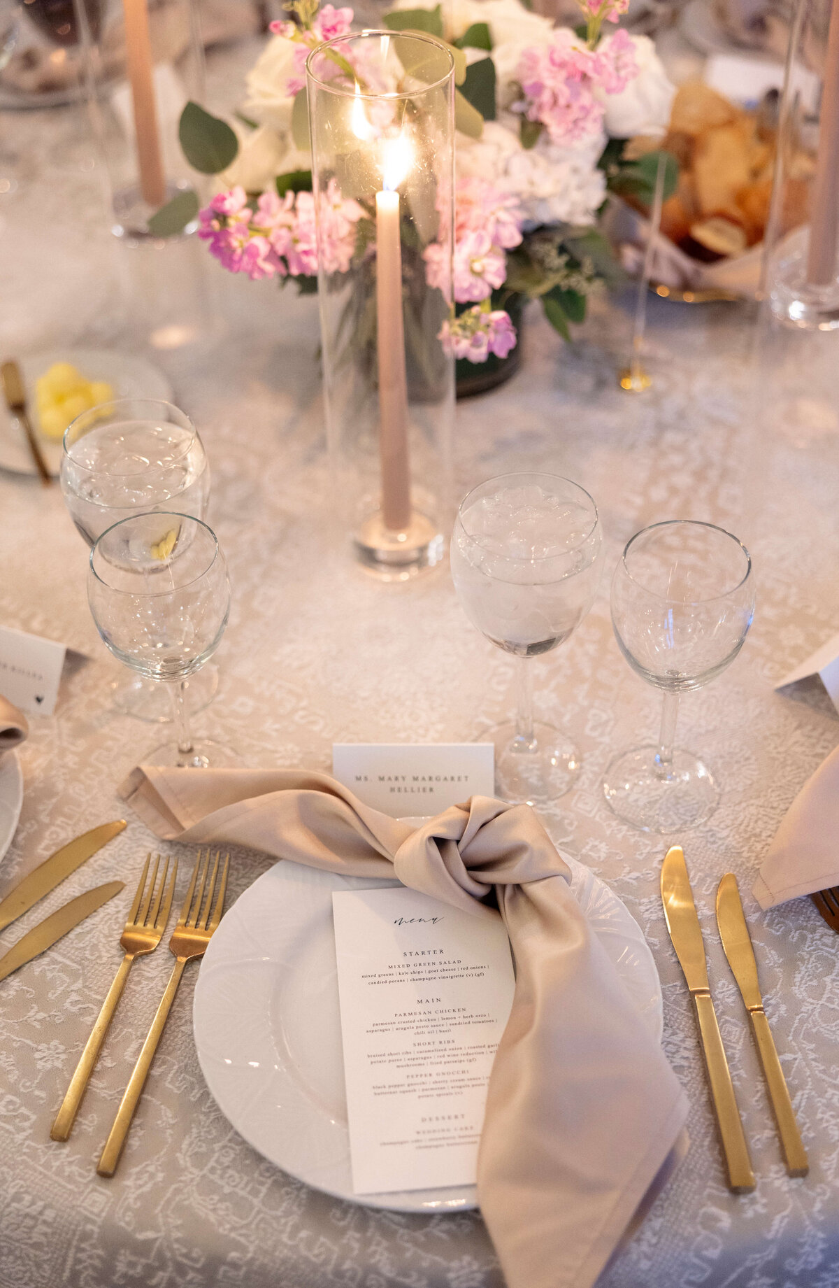 A sophisticated table setting featuring a white plate with a beige napkin, gold cutlery, wine glasses, and a menu. A tall candle and a floral centerpiece with pink and white flowers add elegance to the scene.