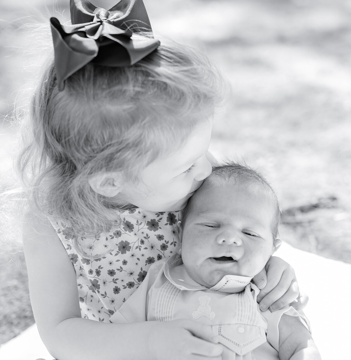 A proud big sister with curly red hair and a bright blue bow grins while holding her newborn baby sibling in a soft, natural outdoor setting. This sweet sibling moment captures the joy and tenderness of welcoming a new baby, reflecting the light and heartfelt style of a Birmingham newborn photographer.