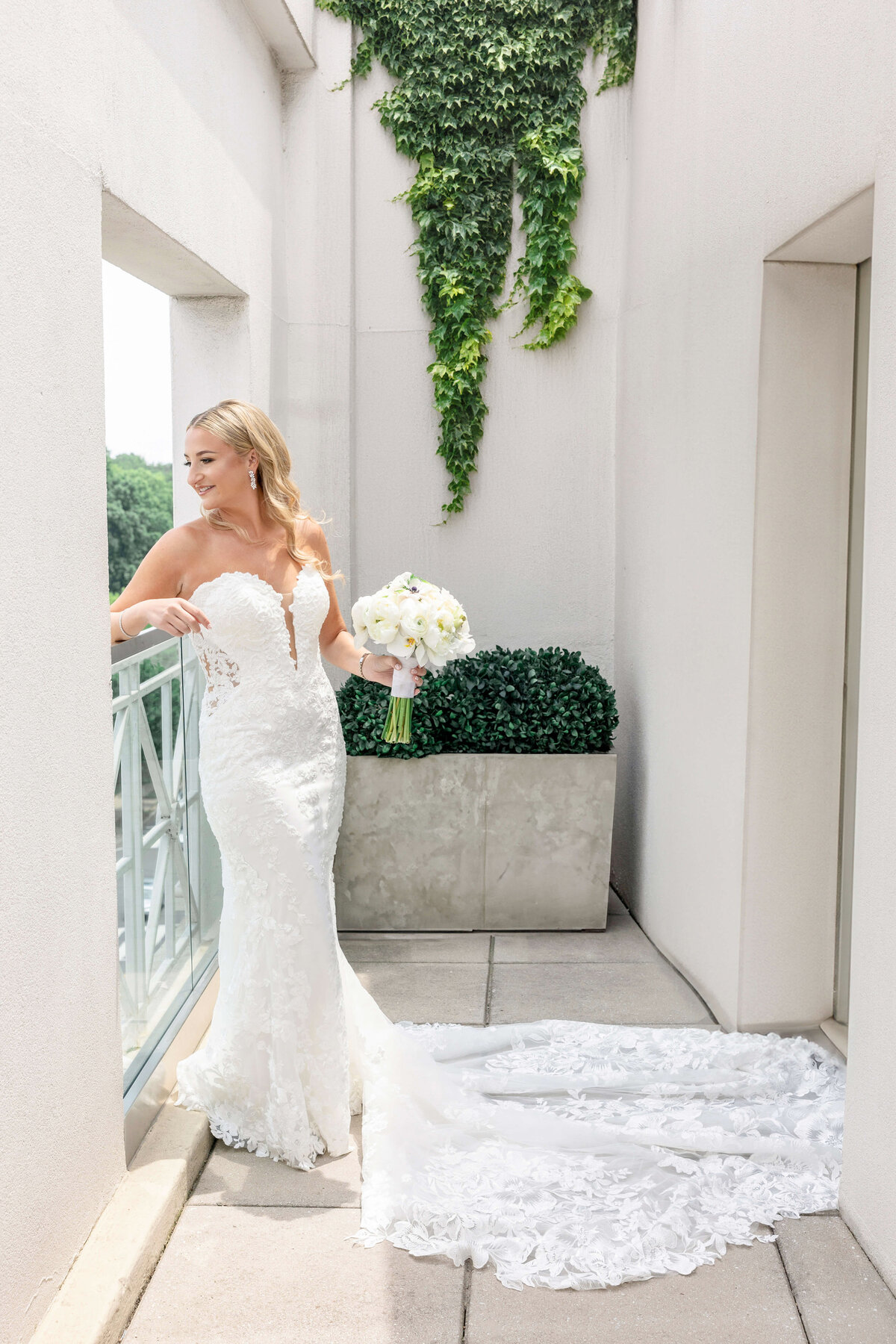 A bride in a white lace wedding gown stands on a balcony, holding a bouquet of white flowers. Green ivy decorates the white wall behind her. The train of her dress is spread out elegantly on the floor.