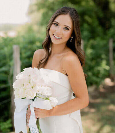 Bride with long brunette waves and glowing makeup smiling outdoors, showcasing soft bridal hair and makeup by a professional makeup artist with full wedding day services