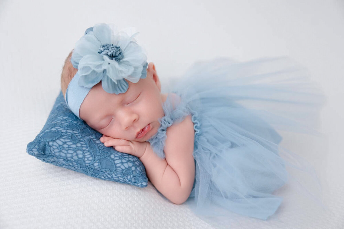 Newborn girl posed on a white blanket and wearing a blue laced dress with a blue-flowered bow and lying on a blue-laced pillow.