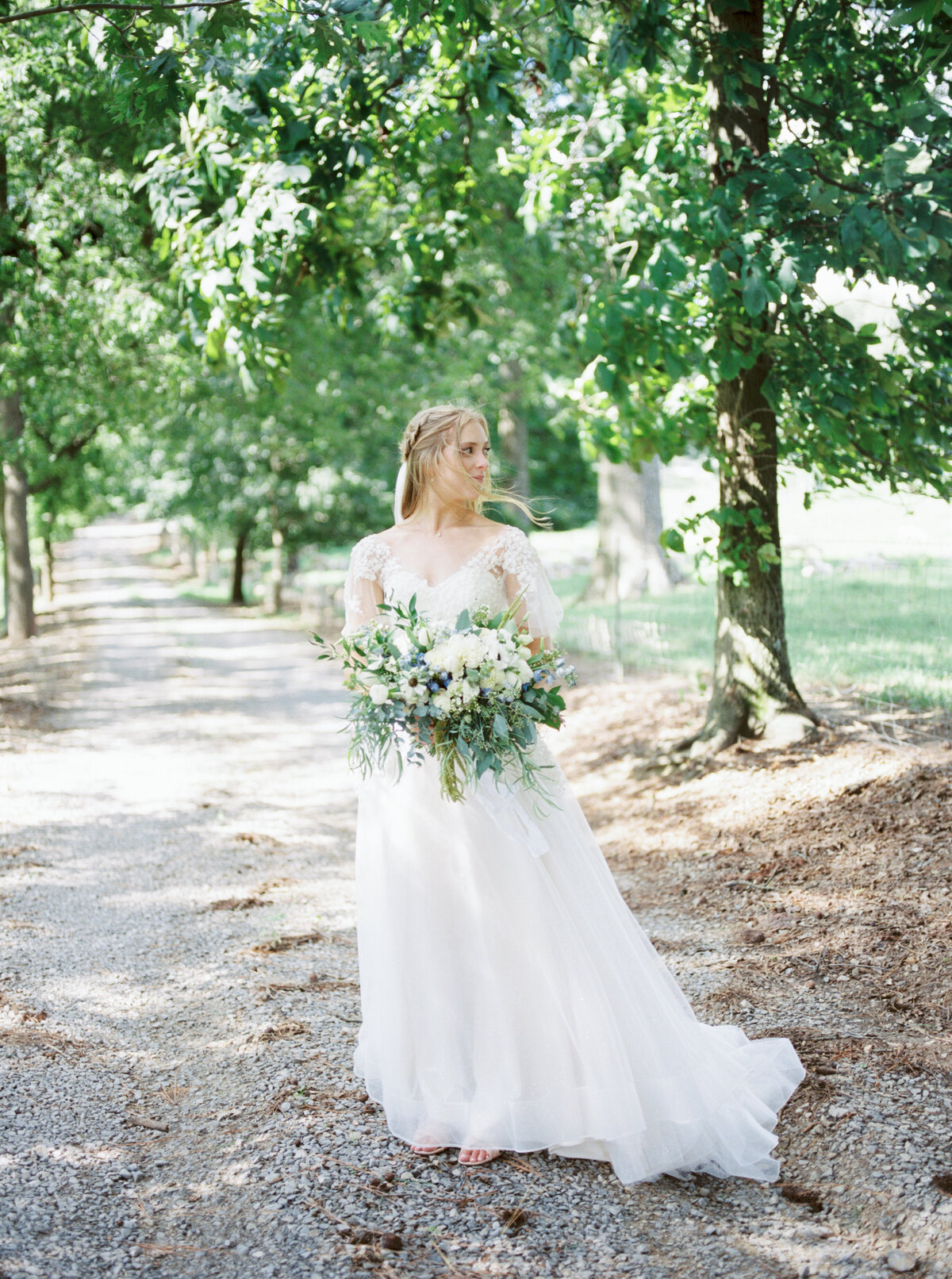 A bride stands on a gravel path with her bouquet while wind blows through her hair by Birmingham wedding photographer, Kelsey Dawn Photography