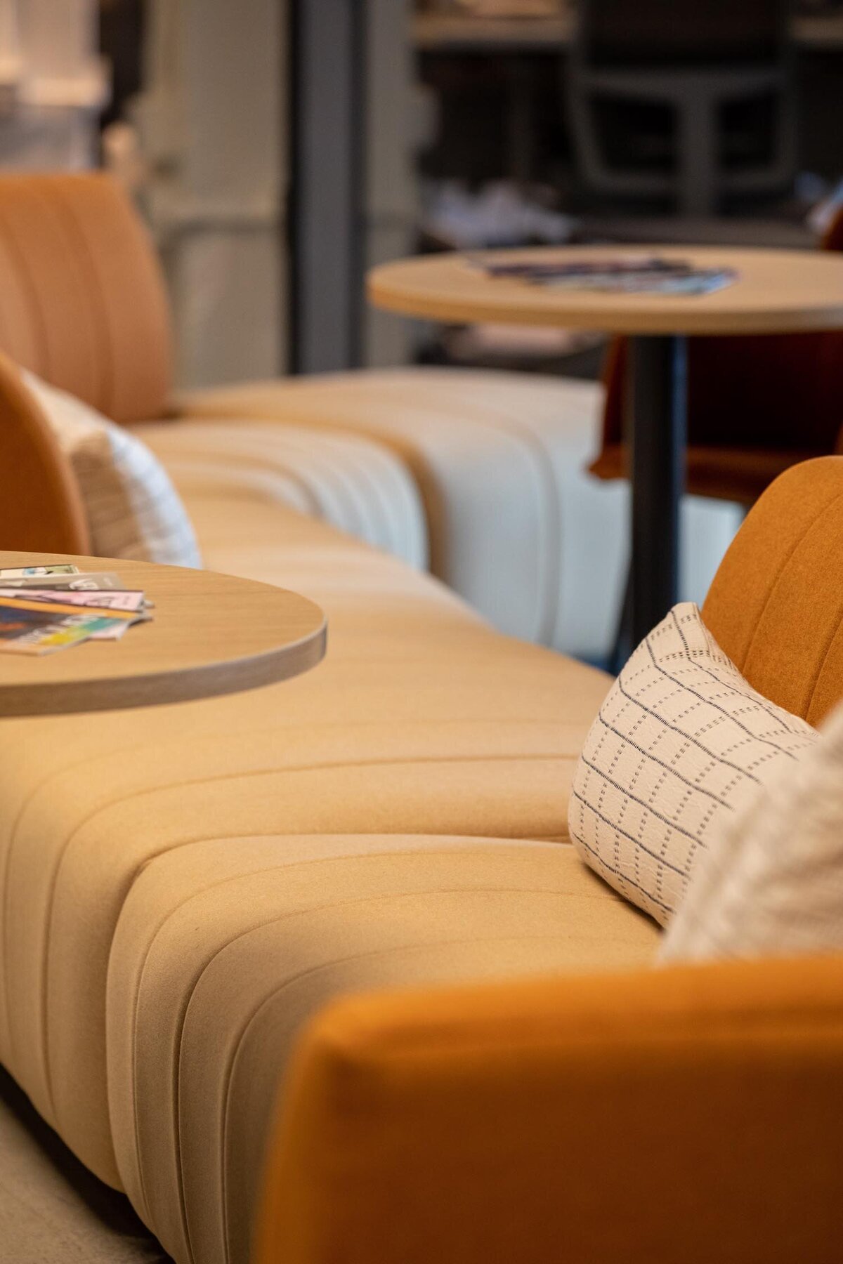 Upholstered beige lounge bench with striped cushions and round tables arranged throughout a communal space.