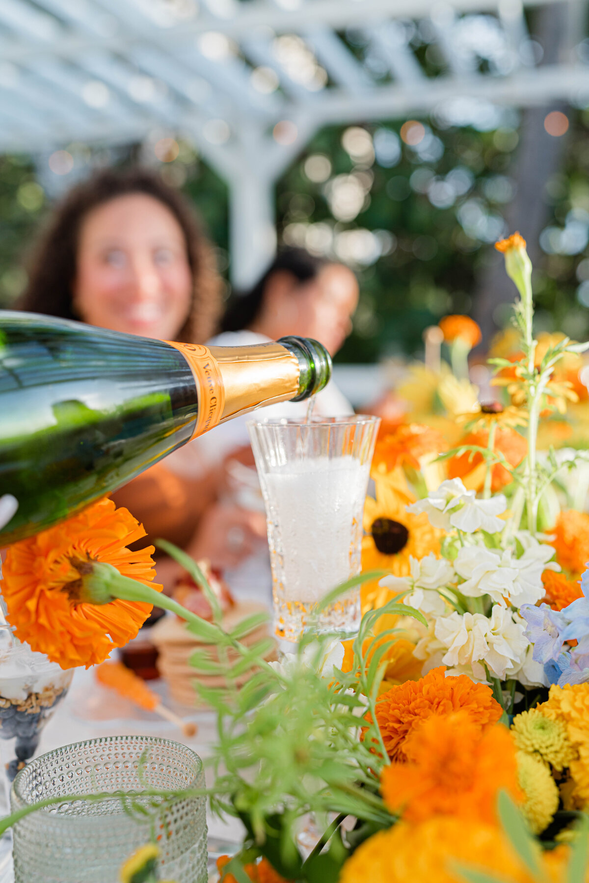  white, orange, blue and green floral table runner with a girl blurred in the background and a bottle of champagne being poured into a glass in focus