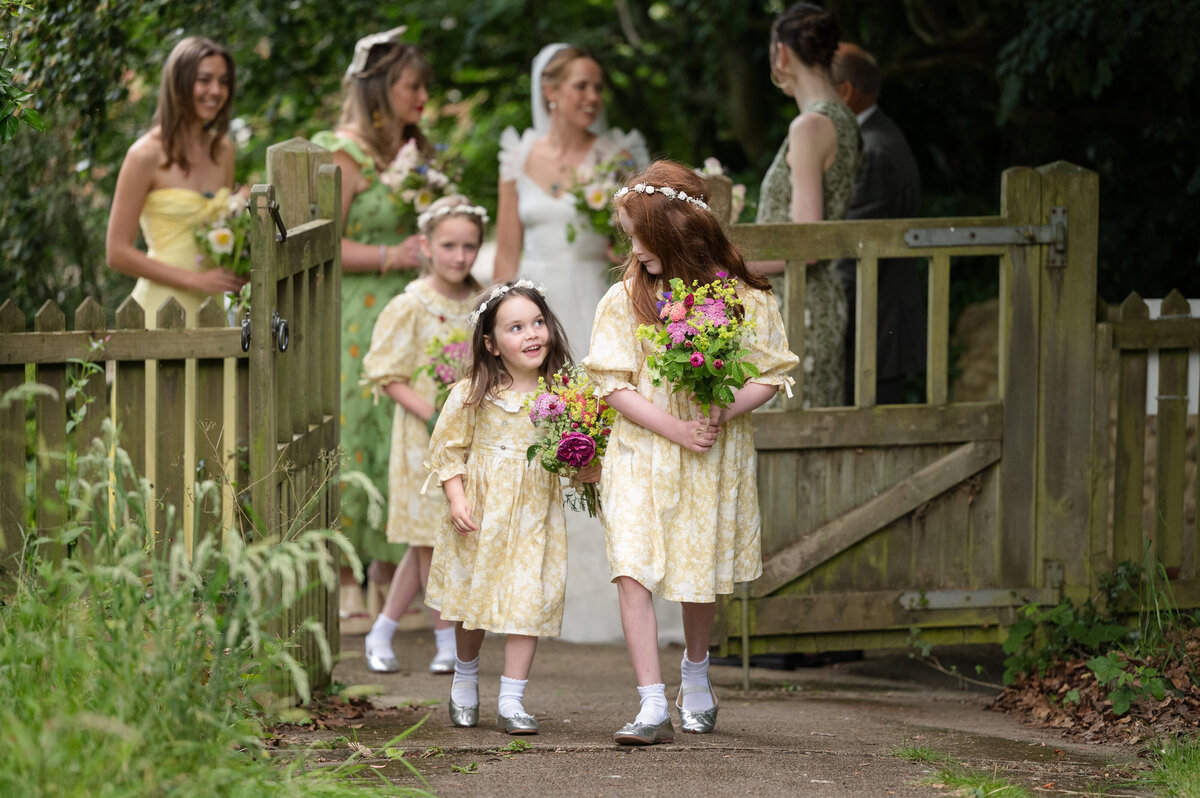 Three flower girls in yellow dresses walking through a wooden church gate