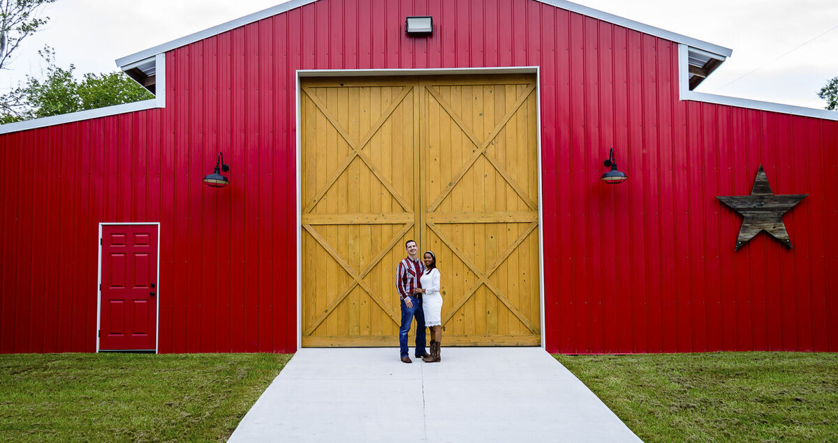 Couple poses for engagement photos in front of a red bard with giant barn doors.