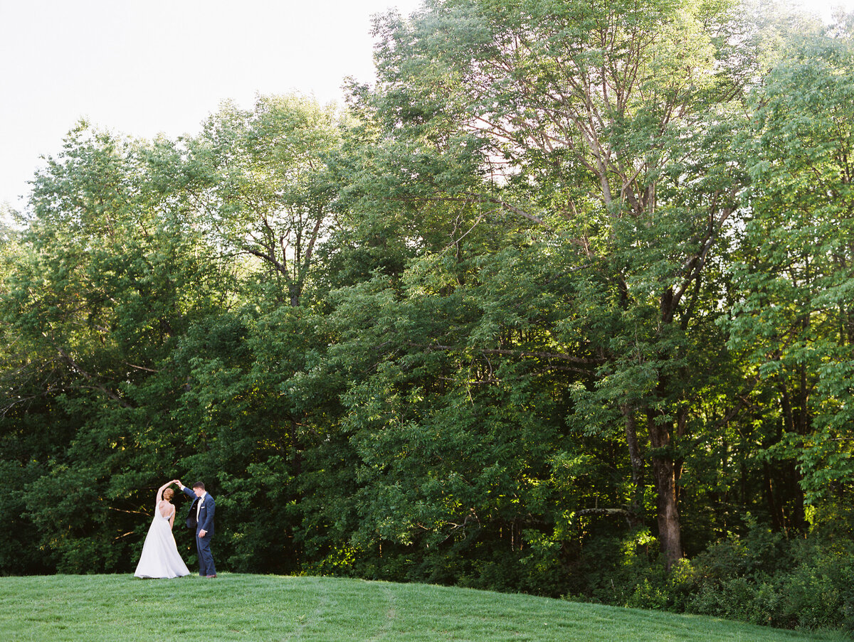 Faraway photo of a bride and groom dancing in a grassy field with trees behind them