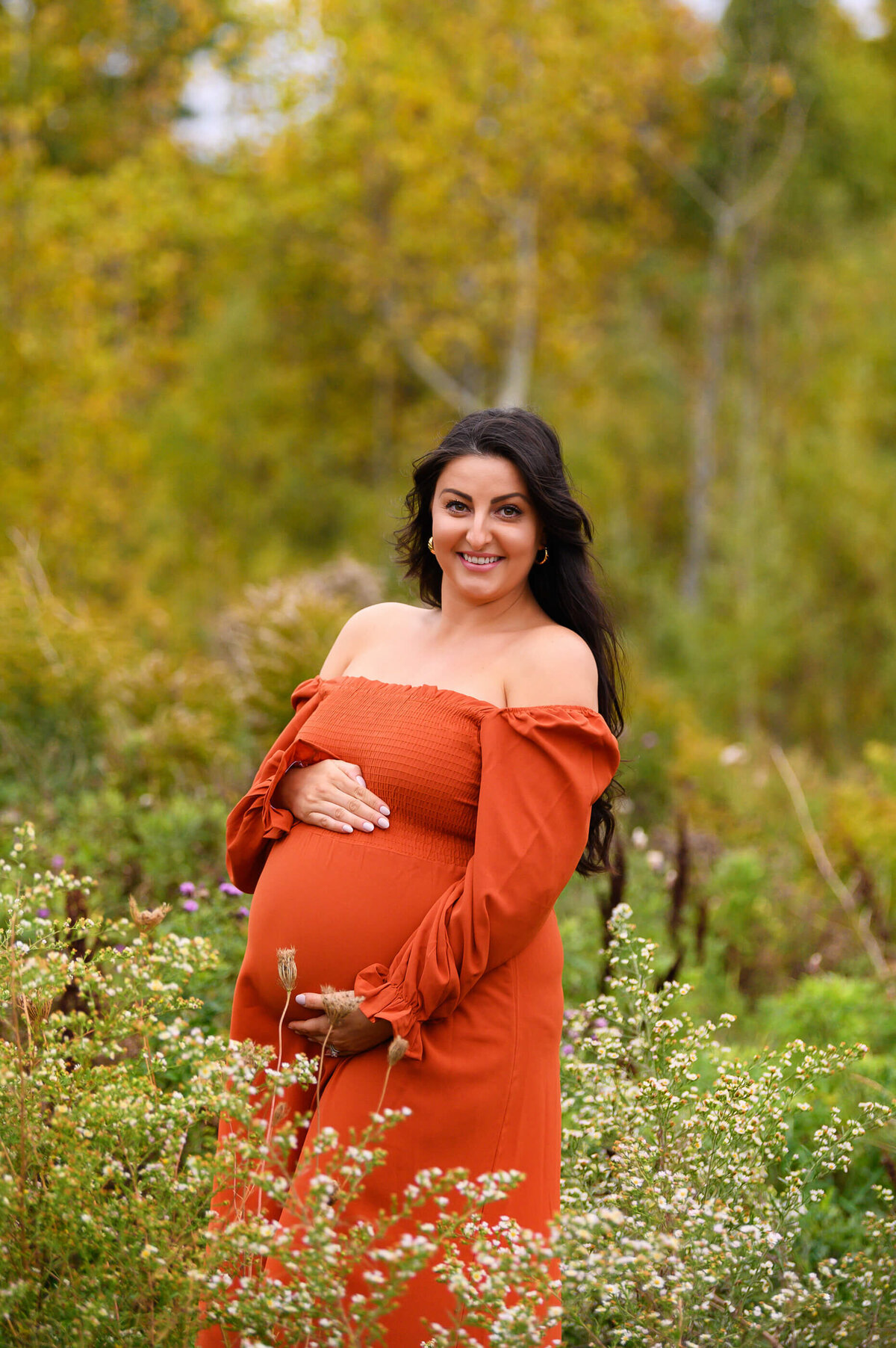 brown-haired pregnant woman wearing a orange maternity dress cradling her belly for her fall outdoor maternity photography session in Burlington, ON