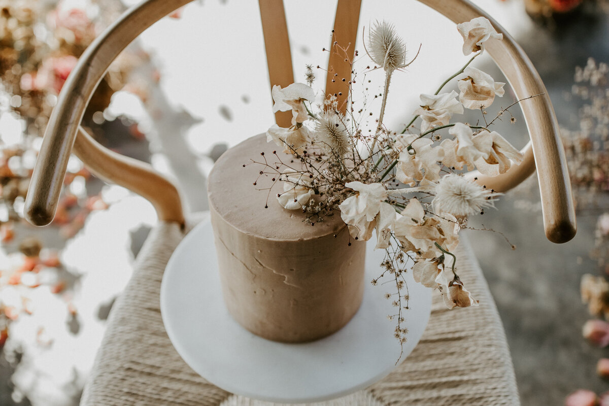 Tan wedding cake with ivory flowers on a white cake stand atop a wishbone chair with flower arrangements in the background.