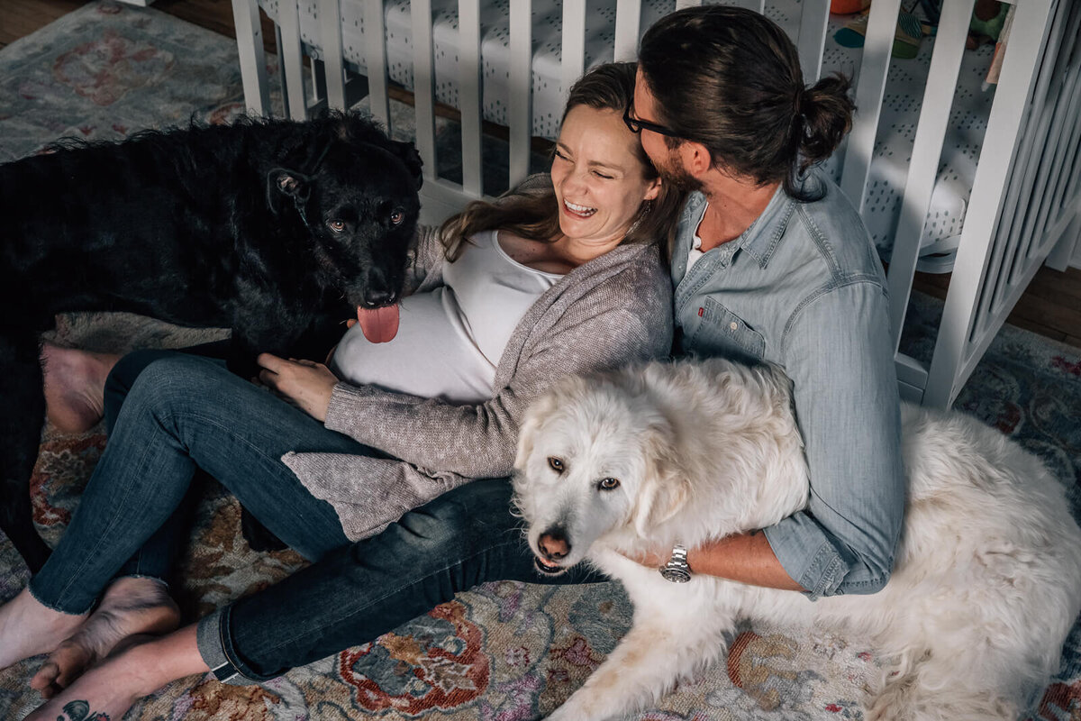 A pregnant mother sits on the floor in front of her baby's crib with her husband and two dogs during an in home maternity photography session with Kate Simpson.