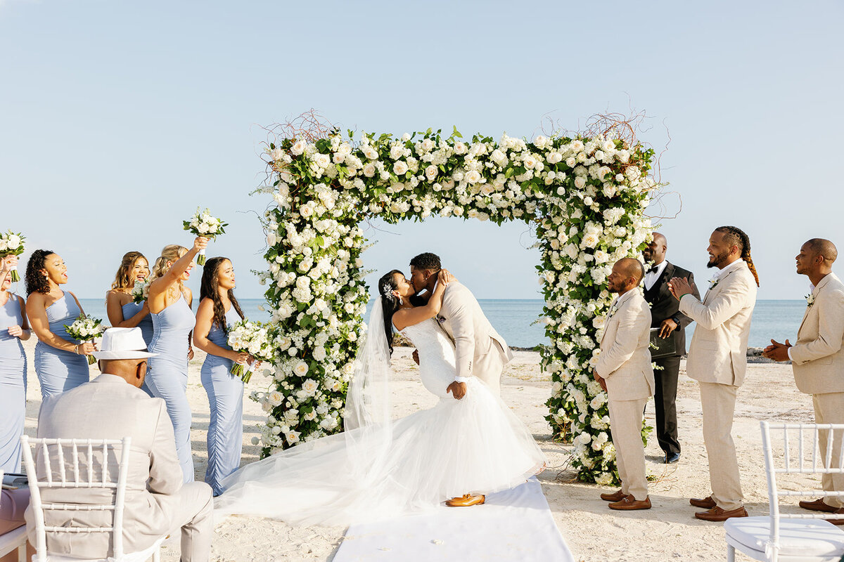 Andressa and Jonathan share a romantic first kiss as newlyweds beneath a lush floral arch at their beachfront wedding in Table Key, Islamorada. Captured by Claudia Amalia, Islamorada wedding photographer.